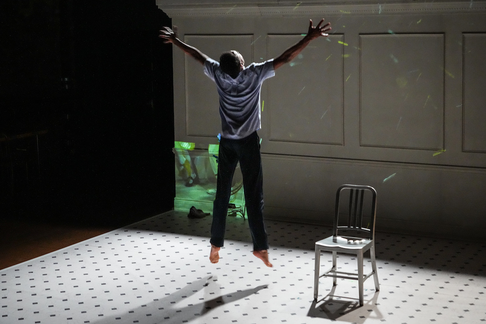 a black man in jeans and light blue short sleeved shirt jumps up arms outstreched as a film is played  on the wall to his leftt